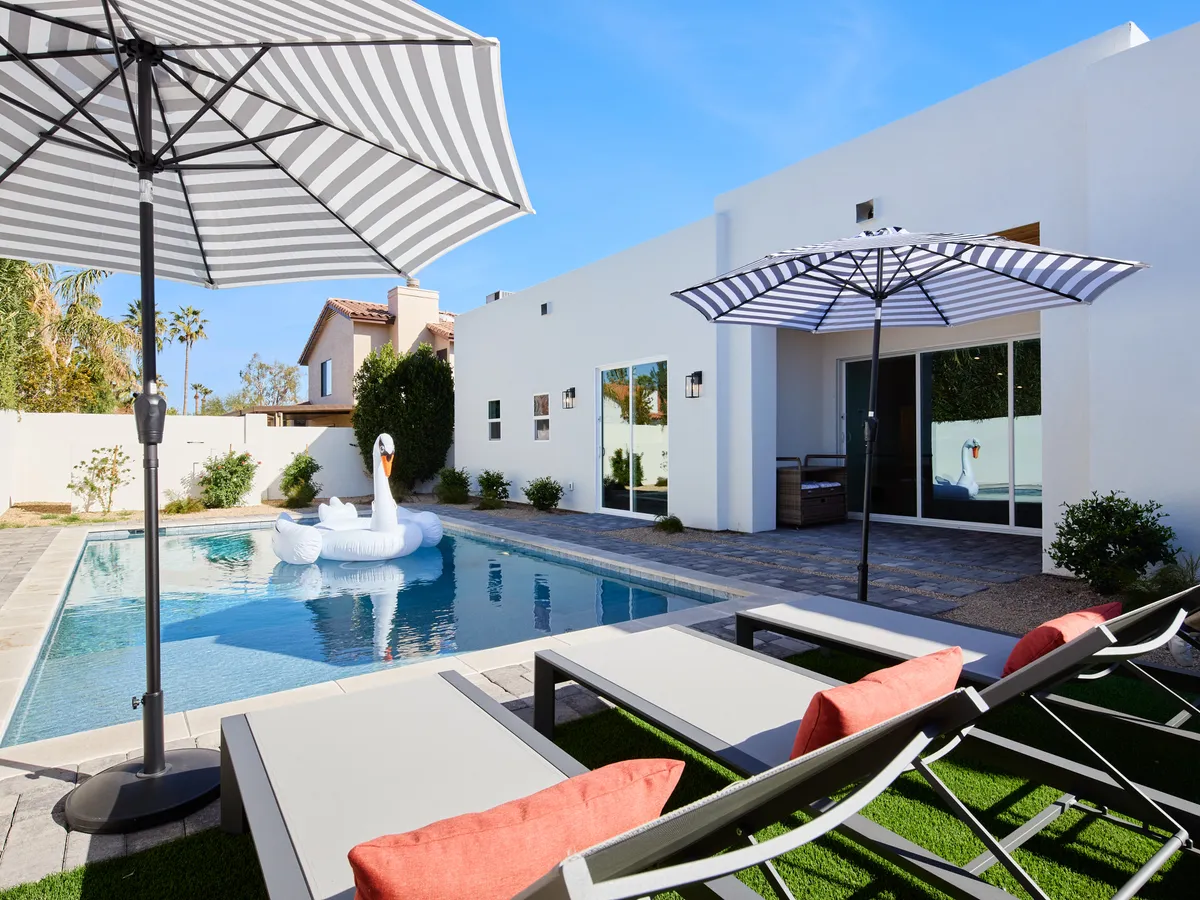 A sun-drenched poolside view featuring sun loungers, umbrellas for ample shade, and a whimsical swan float. The setting evokes lazy afternoons, refreshing swims, and socializing with friends or family.