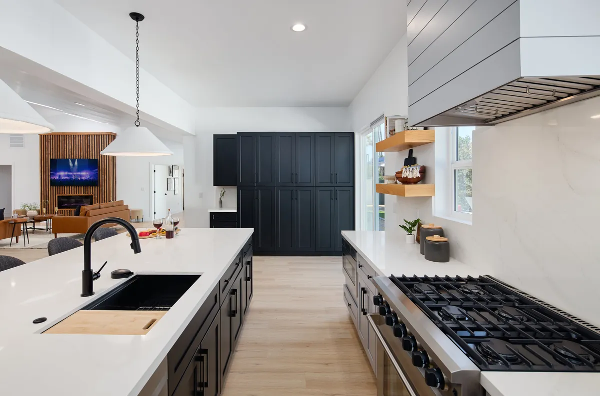 A sleek, modern kitchen with a large central island, black cabinetry, stainless steel appliances, open shelving, and stylish lighting fixtures. The space feels welcoming and perfectly set up for group cooking or entertaining.
