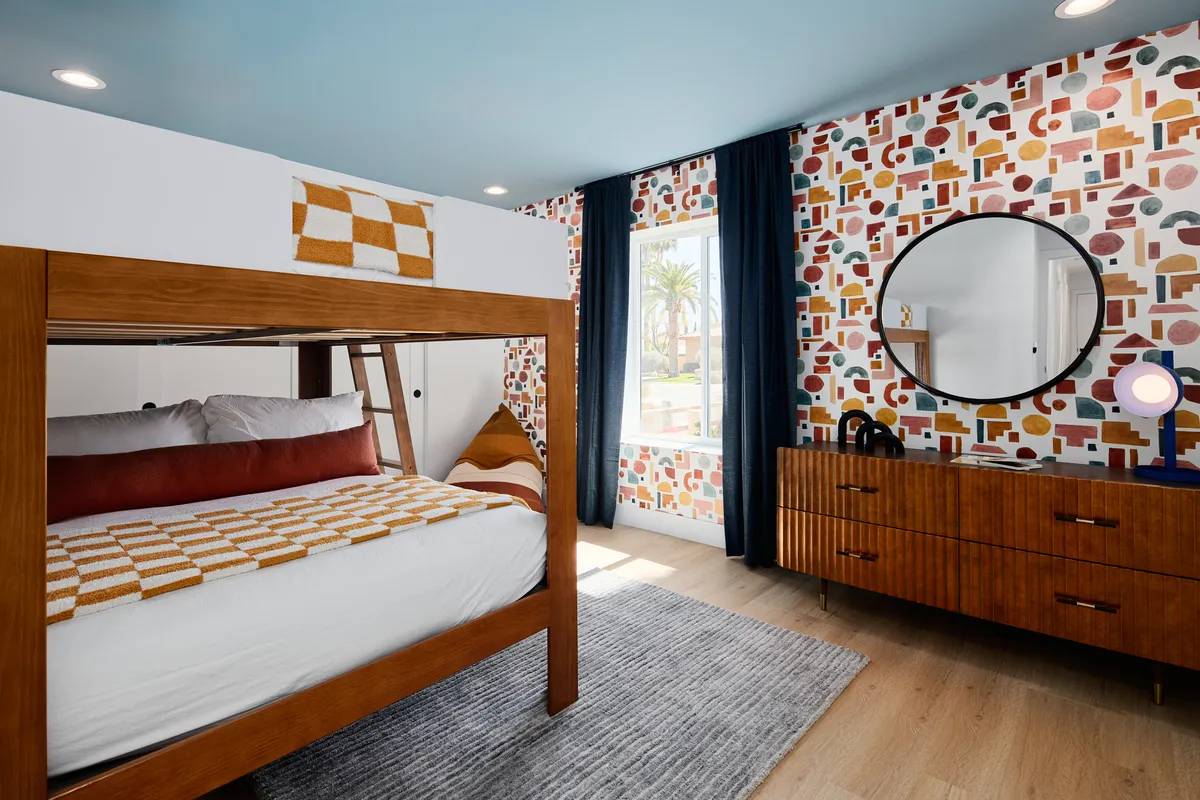 A cheerful bunk room with a bunk bed, colorful patterned wallpaper, a round mirror, and a wooden dresser. The decor is playful and inviting, perfect for children or groups to enjoy sleepovers and stories together.
