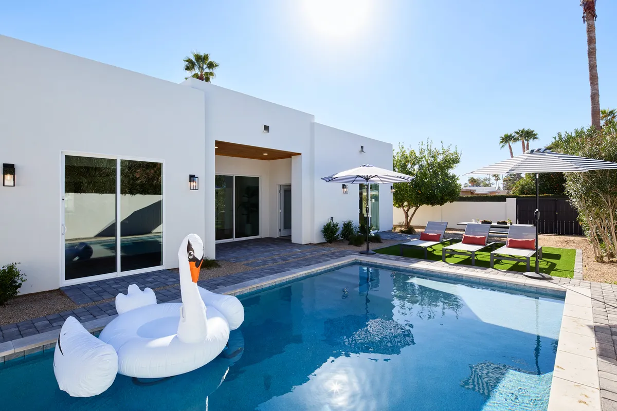 The image showcases a vibrant backyard scene with a sparkling blue swimming pool at its center, complete with an inviting inflatable swan floating on the water. Surrounding the pool are stylish outdoor seating areas shaded by sun umbrellas, all set against a backdrop of tall palm trees, creating a lush, resort-like atmosphere. The scene suggests opportunities for swimming, lounging with a drink, hosting playful summer gatherings, and unwinding under the Arizona sun.