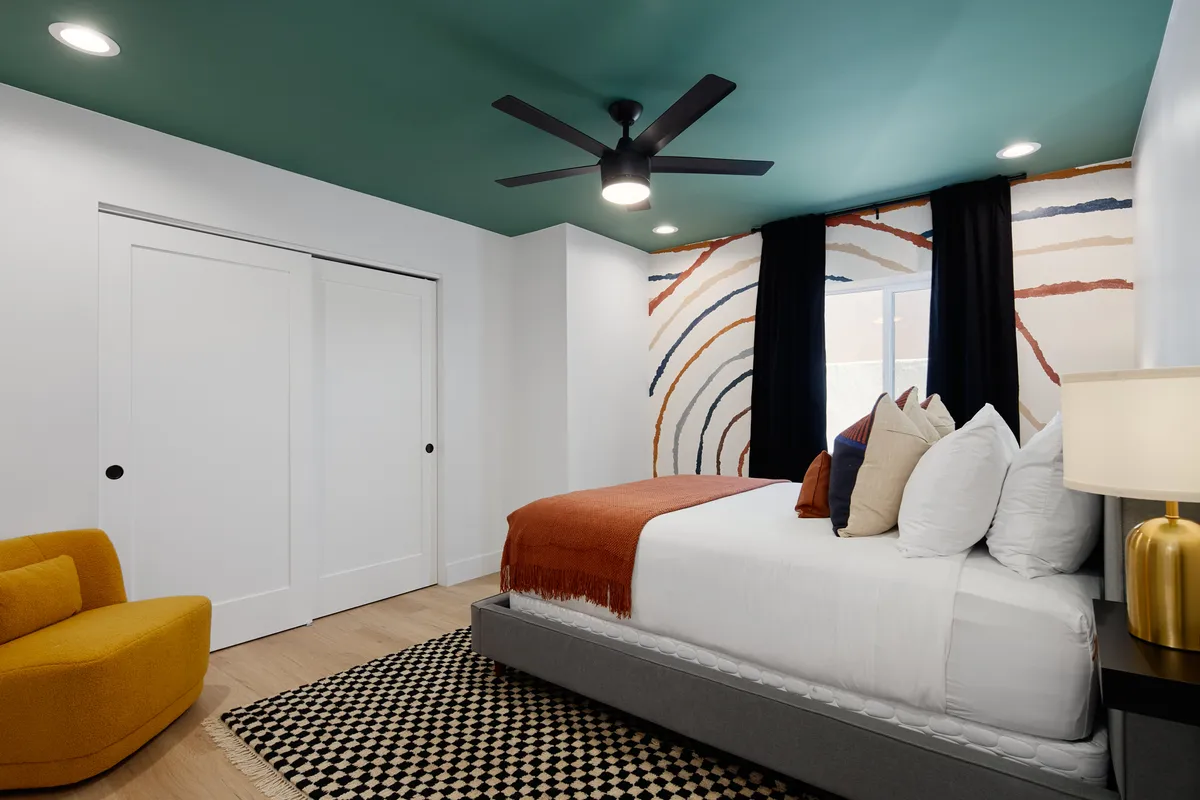 A lively bedroom featuring a bed with decorative pillows, a colorful accent wall, a modern ceiling fan, and a bold yellow accent chair. The vibrant decor and inviting light create a cheerful ambiance.