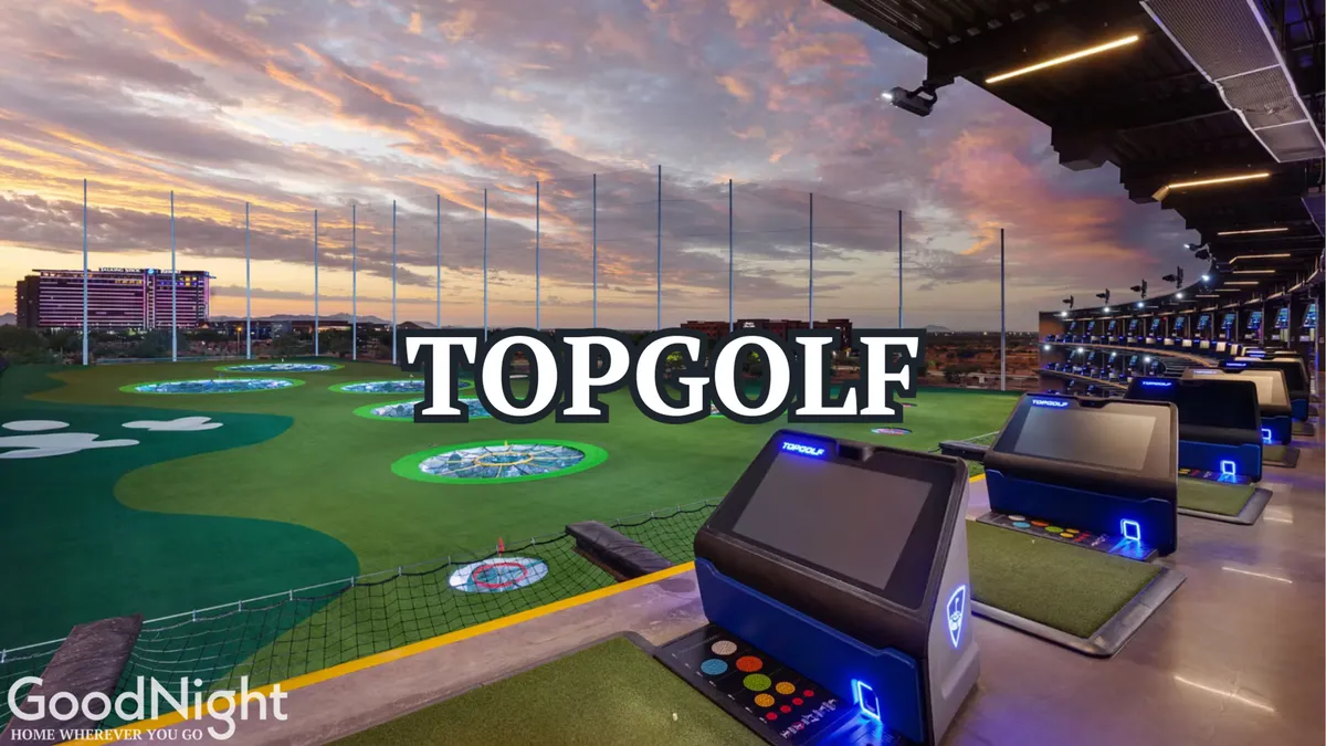 The image shows the lively atmosphere at a Topgolf-style driving range, with multiple golf bays overlooking a skyline and illuminated LED screens. Groups are seen enjoying food and drinks while competing in interactive golf games against a sunset backdrop.