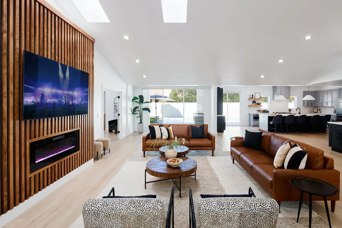 A contemporary, open great room featuring leather sofas, a large TV mounted above a modern fireplace, a stylish coffee table, decorative pillows, and an indoor plant. The space is airy and inviting, with thoughtful decor and easy flow for mingling and entertainment.