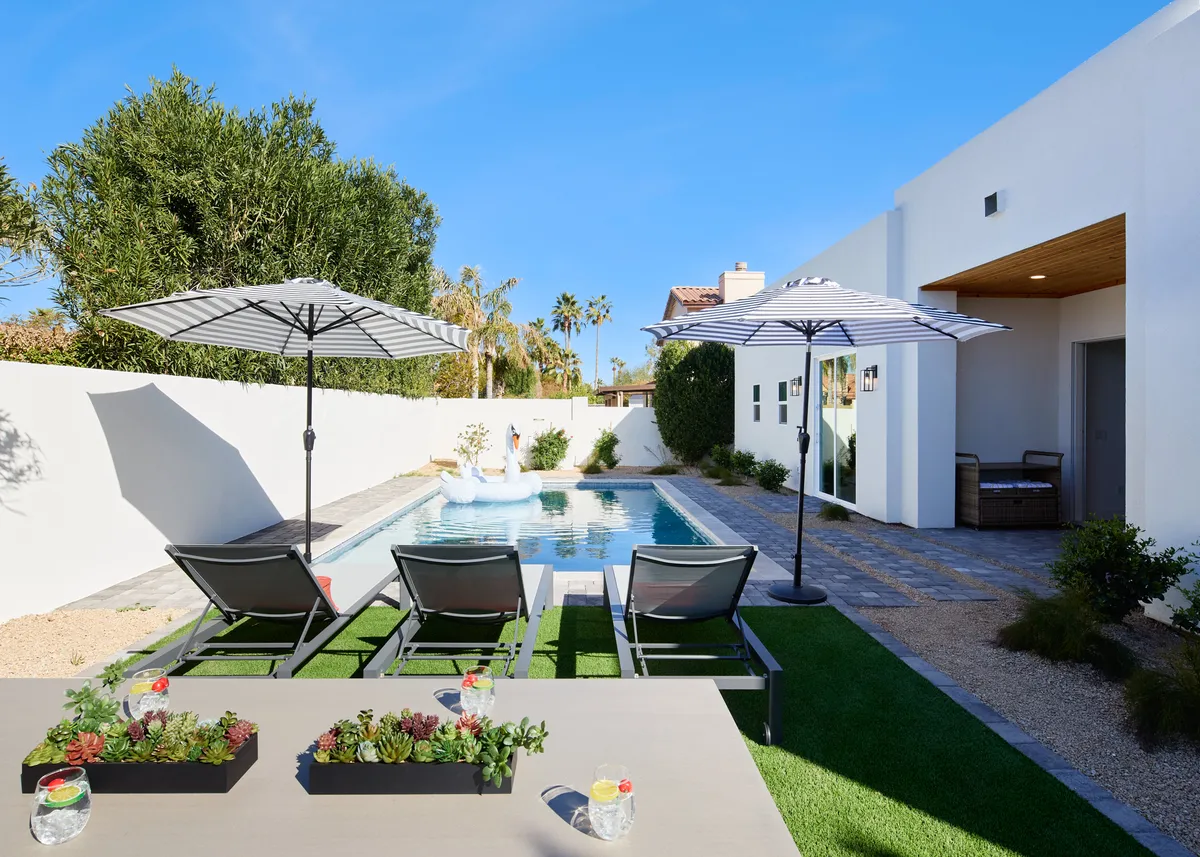 A beautifully landscaped backyard featuring a sparkling swimming pool surrounded by lounge chairs, umbrellas for shade, and succulent planters. The area appears tranquil and inviting, ideal for relaxing, swimming, or social gatherings.