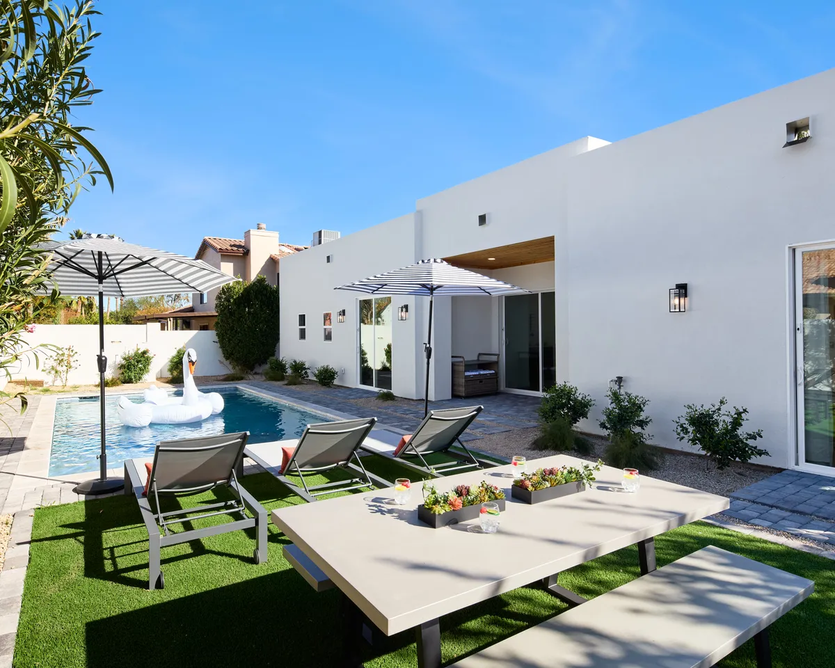 A cheerful backyard pool scene with a large swan float, outdoor dining table shaded by an umbrella, lounge chairs, and lush greenery. The setup highlights areas for both sunbathing and al fresco dining, all in a private, landscaped environment.